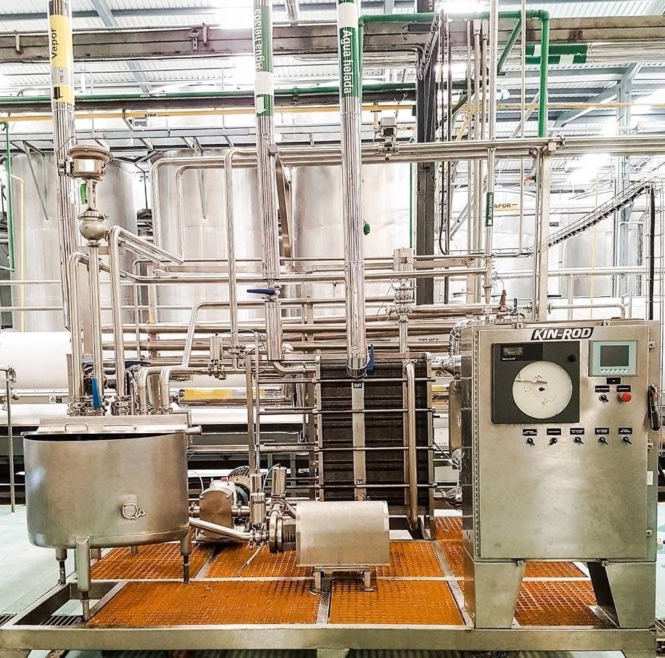 Pasteurizador HTST KIN-ROD instalado en planta industrial con tablero de control y logotipo KIN-ROD visibles, tuberías de vapor y agua helada, tanque de proceso y piso industrial naranja